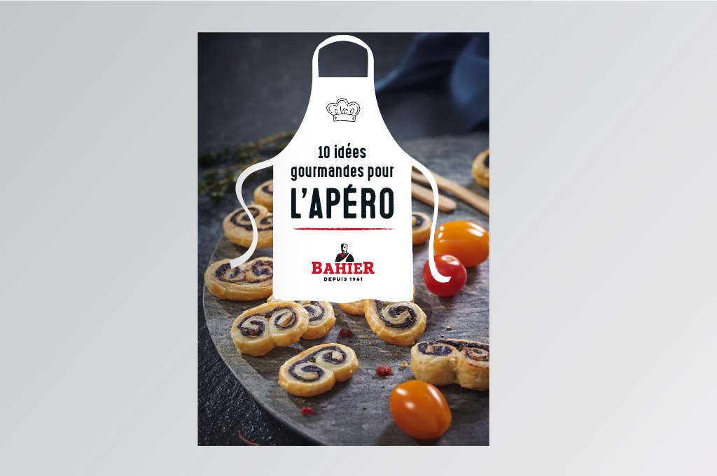 Couverture d’un livret de recettes Bahier de 16 pages consacré à l’apéritif. Sur un fond sombre et élégant, une planche en ardoise présente des feuilletés apéritifs en forme de spirales garnies, accompagnés de tomates cerises rouges et jaunes. Au centre de l’image, une illustration de tablier blanc contient le titre « 10 idées gourmandes pour l’apéro ». En bas du tablier figure le logo Bahier avec la mention « Depuis 1941 ». L’ensemble évoque la convivialité et la gourmandise autour de la charcuterie. Réalisé par l'agence A tribu - Le Mans