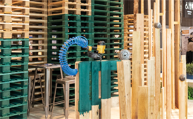 Détail mise en scène fabrication palette neuve avec un pistolet à clou en équilibre sur une palette et une composition de planches verticales sur lesquelles des rouleaux de clous sont disposés - vue du stand de 100m2 conçu par Atribu pour PGS group, sur le salon all4pack 2022