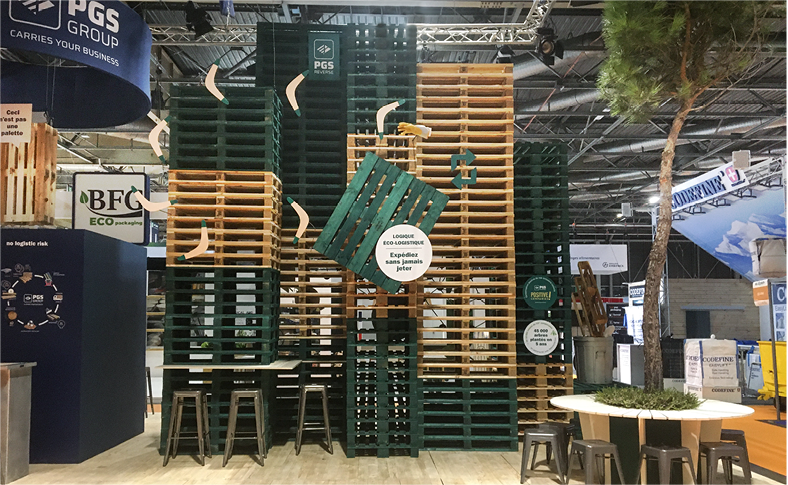 vue du stand de 100m2 conçu par Atribu pour PGS group, sur le salon all4pack 2022 - Mise en scène palette verte suspendue et 7 boomerangs avec une main dans un gant de travail